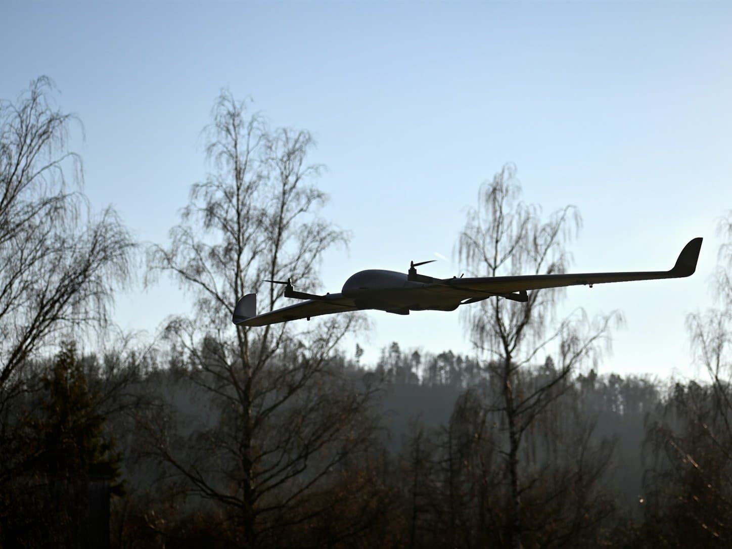 A fixed-wing unmanned aircraft in level flight above a treeline, twin wing-mounted propellers turning